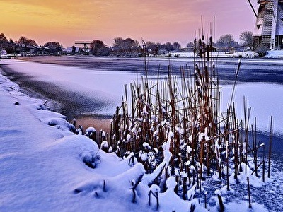 Een windmolen staat in de sneeuw naast een rivier, omgeven door een serene winterlandschap.