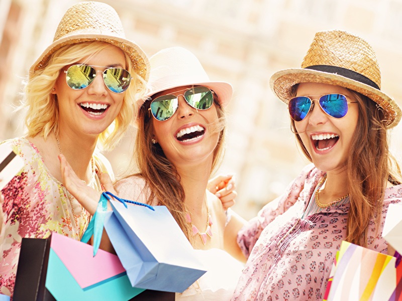 Three happy women wearing sunglasses and straw hats enjoy a shopping day, carrying colorful bags.