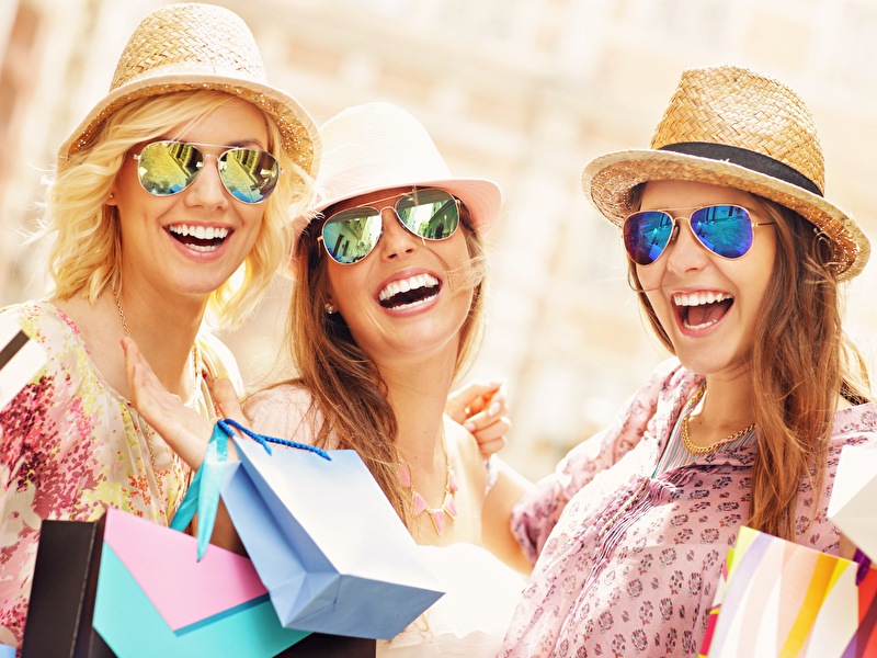 Three happy women wearing sunglasses and straw hats enjoy a shopping day, carrying colorful bags.