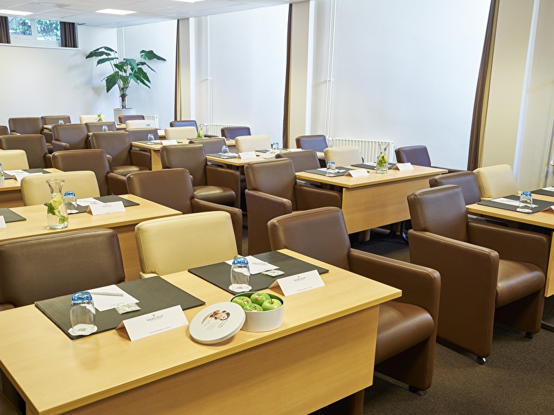 Meeting room with a modern setup, featuring comfortable chairs, wooden tables with notepads, water glasses, and a bowl of green apples.